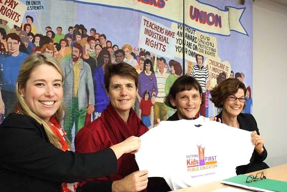 Four women hold upo a t-shirt with a slogan about children and education, in front of a union banner.