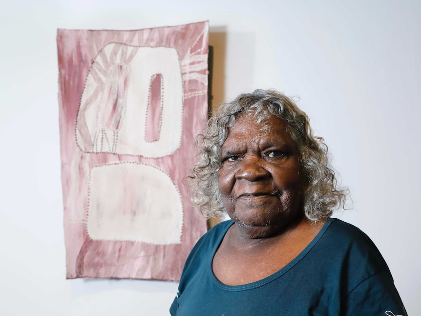 Interior, gallery. Woman with short curly grey hair standing in front of bark painting in pink and white tones.