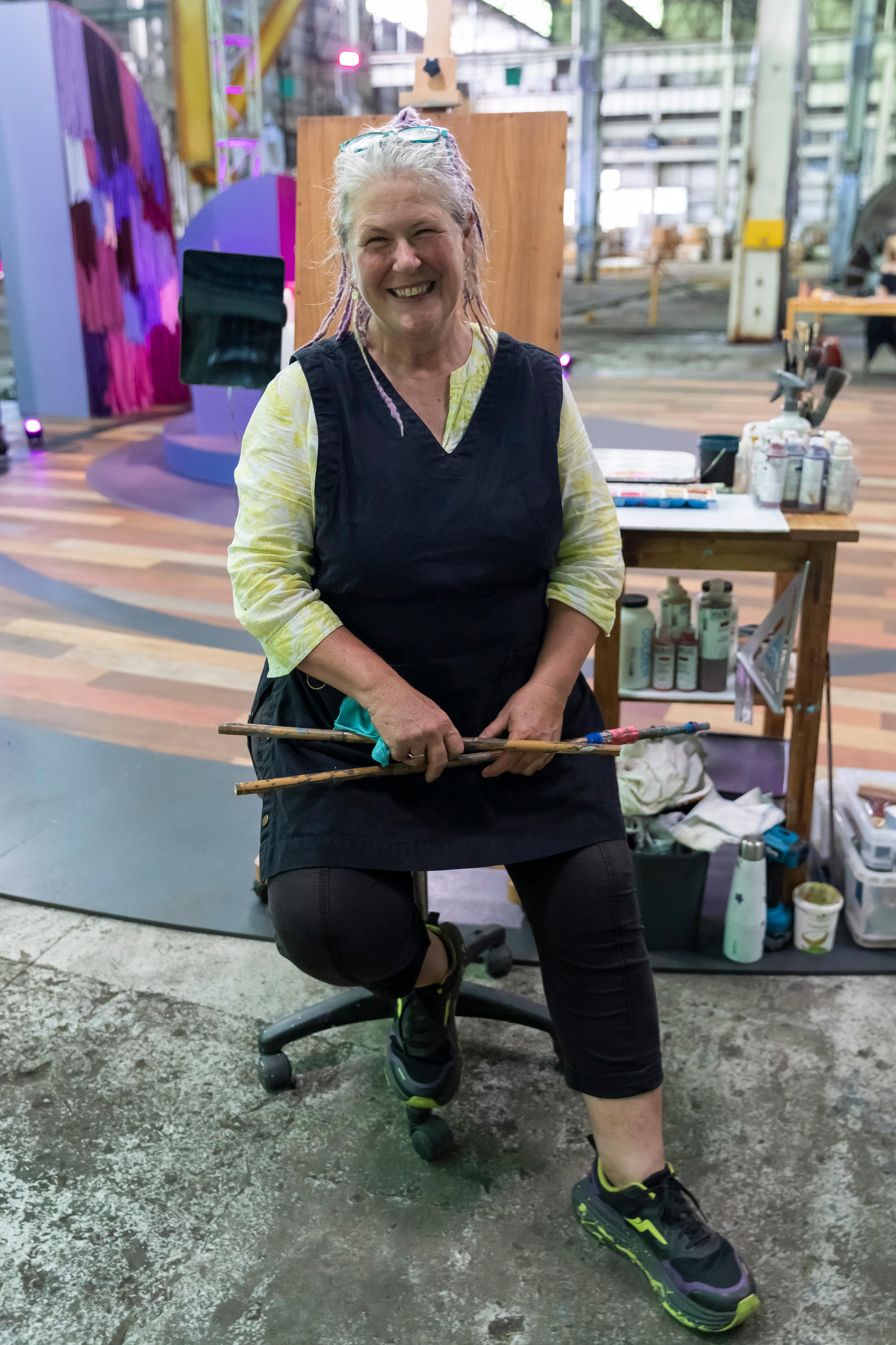 A woman with grey and purple dreadlocks, pulled back, smiles at the camera. She sits in front of a table of art supplies