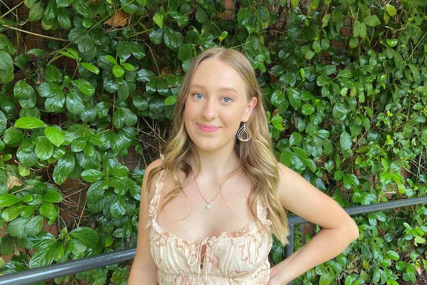 A woman stands in front of a wall of green plants. Her hand is on her waist, her hair is down and she wears a dressy top.