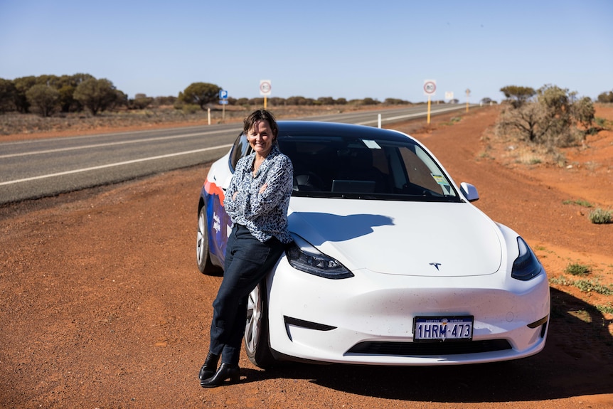 A woman leaning on the bonnet of an electric vehicle alongside a remote highway.  