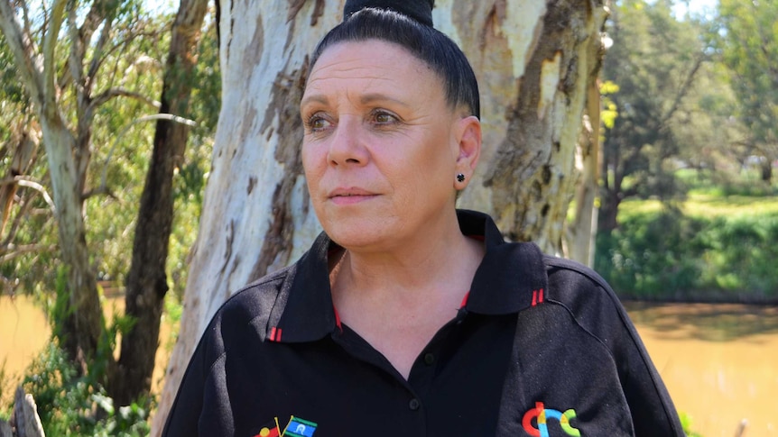 An Aboriginal woman looks into the distance away from the camera with trees and a river behind her