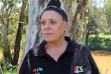 An Aboriginal woman standing in front of a large eucalypt looks into the distance away.