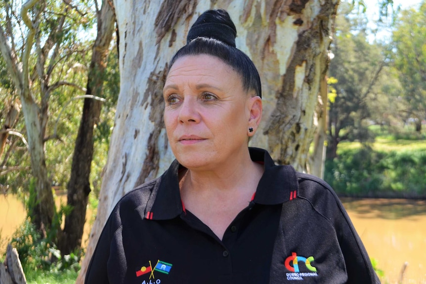 An Aboriginal woman looks into the distance away from the camera with trees and a river behind her