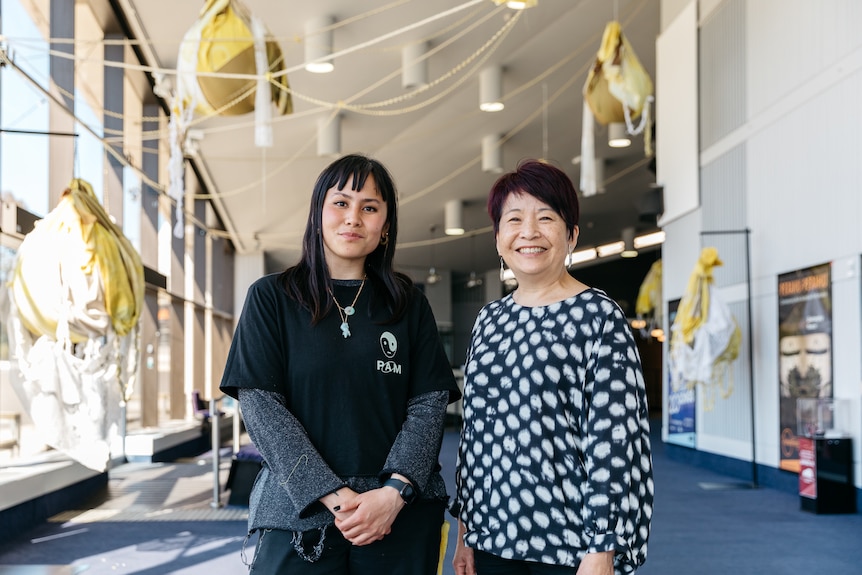 Two Asian Australian women, one in her 30s, the other in her 60s, stand smiling together in a theatre foyer
