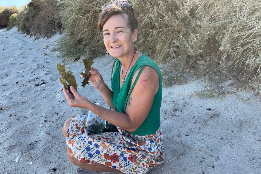 Woman kneels on beach sand displaying two sections of dried seaweed.