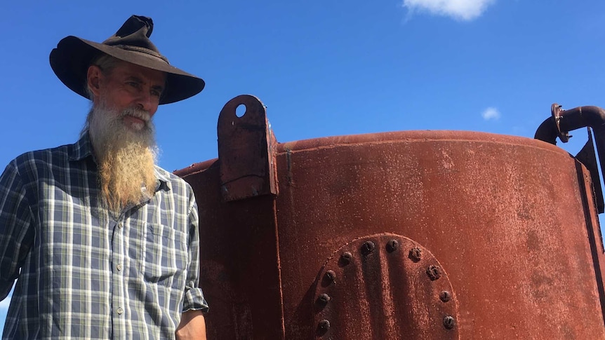 David Bauer with long beard and wizard hat stands next to red rusted tank with sky behind him
