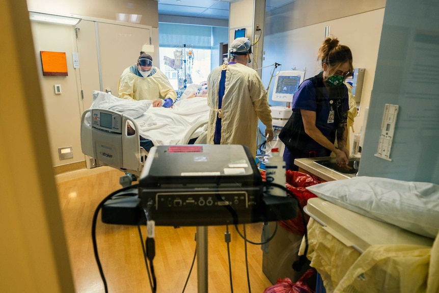Three health professionals in full PPE surround a patient on a ventilator