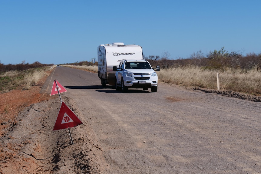 A white car and caravan drive over grey loose gravel section of road. Red warning signs are stuck into the edge of the road.