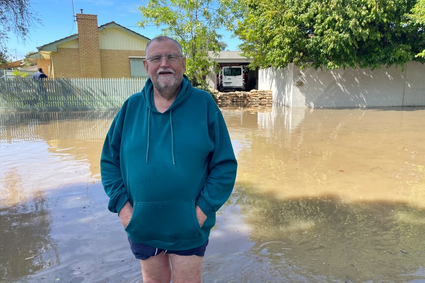 A man stands in front of a flooded street with his home protected by sandbags
