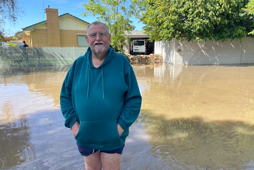 A man stands in front of a flooded street with his home protected by sandbags