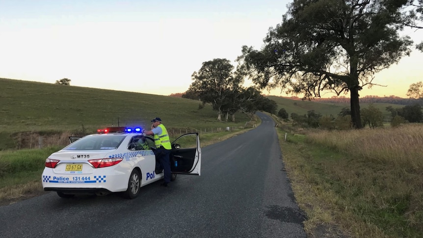 A police officer stands next to his car on a road in the bush.