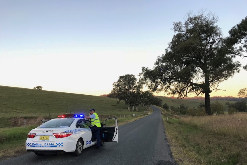 A police officer stands next to his car on a road in the bush.