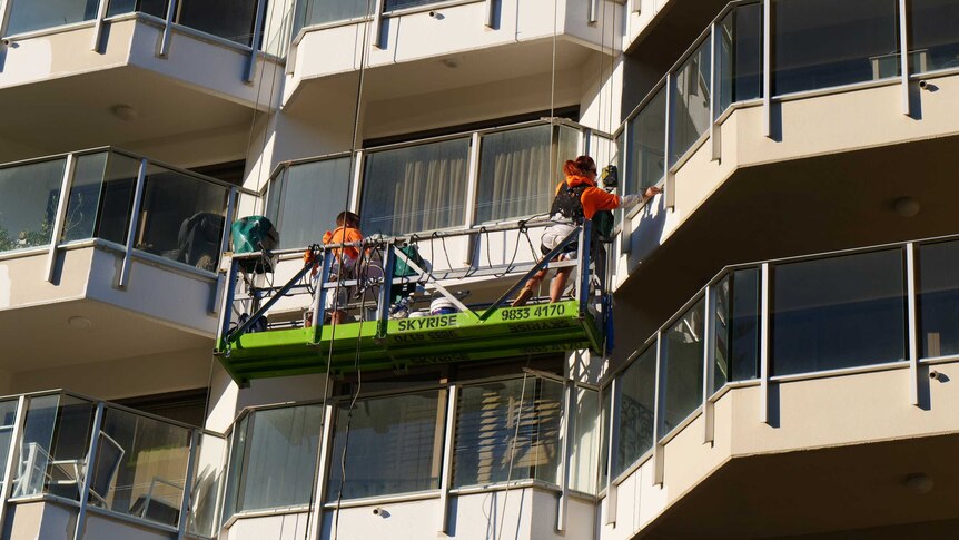 Woman and man with bright orange and white jumper and safety gear strapped on paint highrise from a swing stage