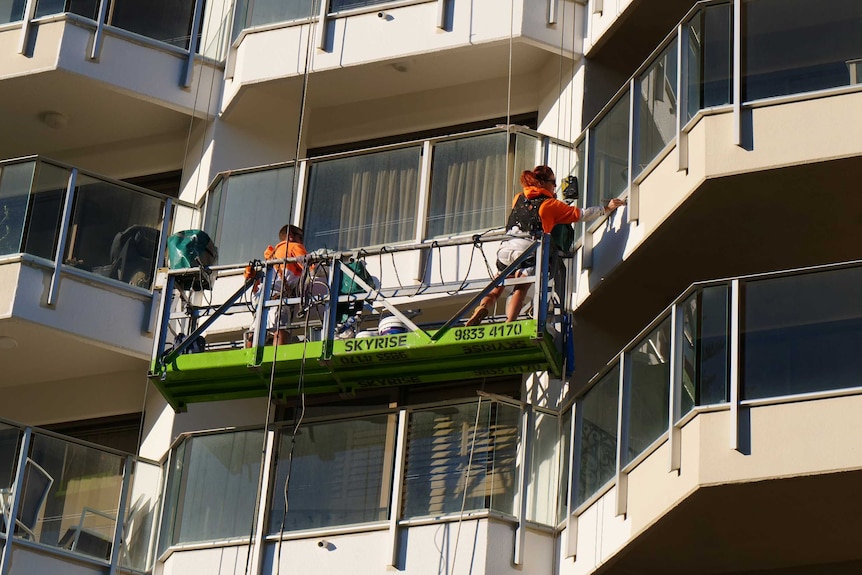 Woman and man with bright orange and white jumper and safety gear strapped on paint high-rise from a swing stage