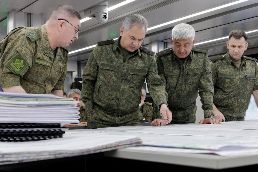 Russian Defence Minister Sergei Shoigu in army gear looking at papers alongside troops.