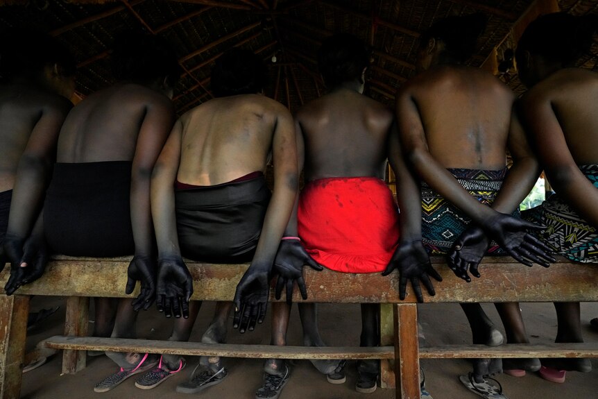 Girls sit on a bench with their backs to the camera, holding their hands dyed black. 