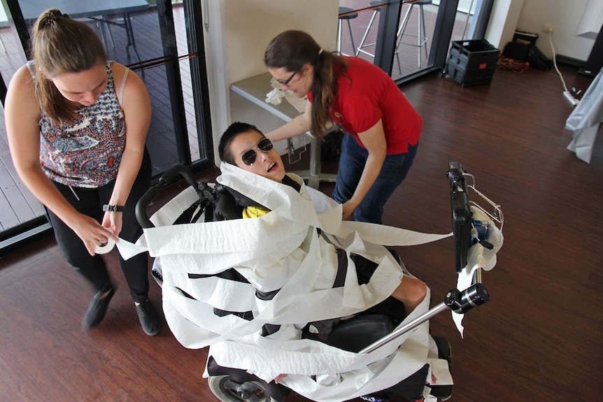 A young man in a wheelchair is wrapped in toilet paper by two women