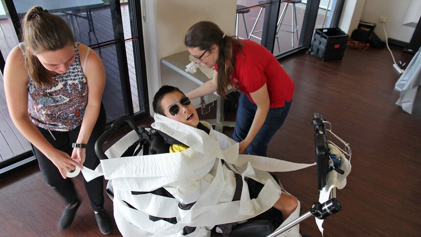 A young man in a wheelchair is wrapped in toilet paper by two women