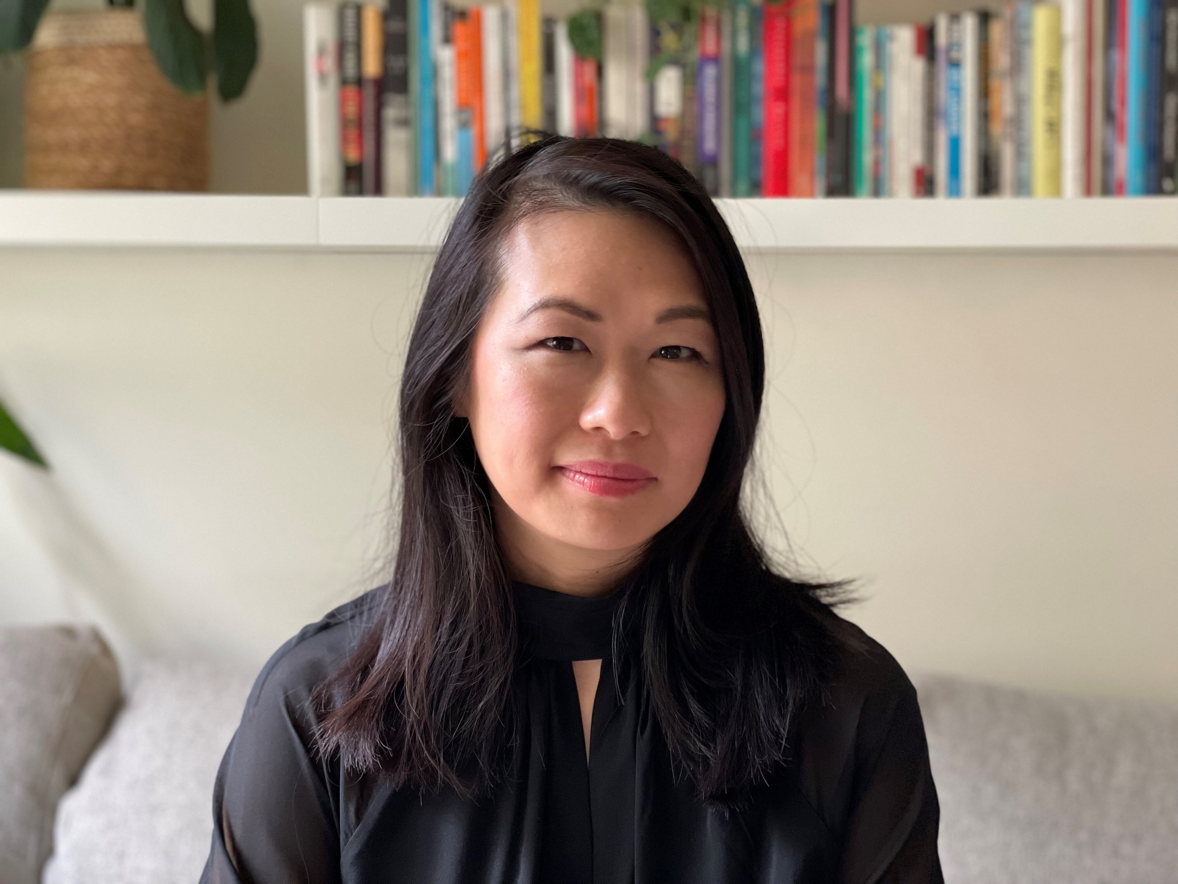 A woman with dark hair sits smiling in front of a bookshelf