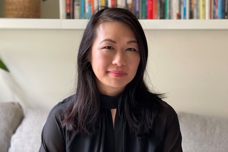 A woman with dark hair sits smiling in front of a bookshelf