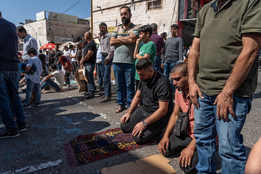 A group of men outside, some kneeling to pray on ornate prayer mats while others stand