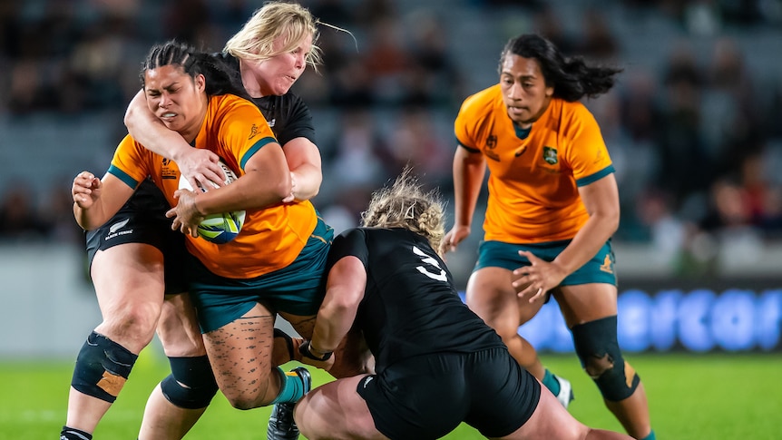 A Wallaroos player holds the ball as she is tackled by two Black Ferns opponents.