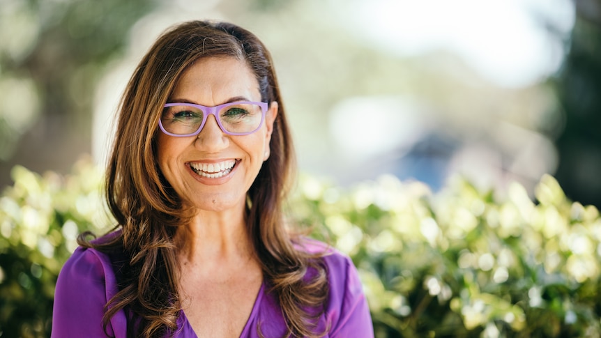 A woman smiles for a portrait wearing purple framed glasses and a purple top.