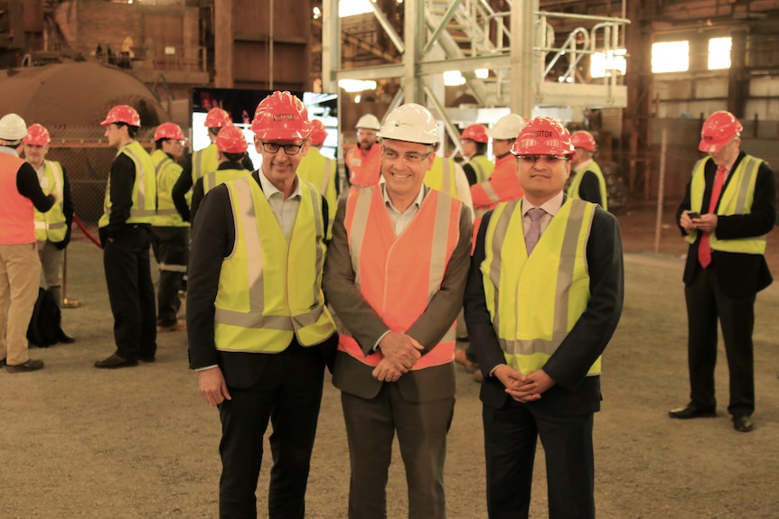Three men stand in high vis and smile inside a large warehouse with people behind them.