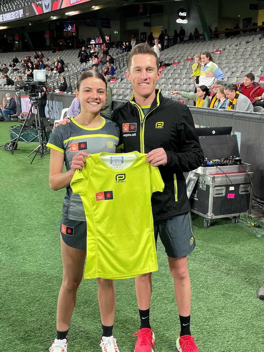 Teenage girl holding umpiring jersey to camera with man at aussie rules game in stadium.