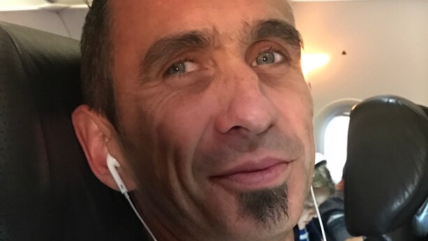 A man nearing middle age, sporting a large "soul patch" beneath his lip, sits on a plane, wearing earbuds.