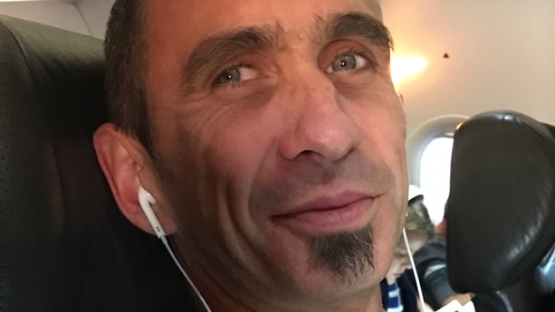 A man nearing middle age, with short dark hair and a "soul patch" under his lip, sits on a plane with earbuds in his ears.