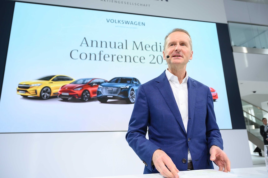 Herbert Diess stands in front of a big screen with different coloured cars on it