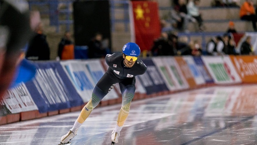 A man wearing ski goggles, ice skate shoes and Australian colours bends forward as he speeds along an ice rink.