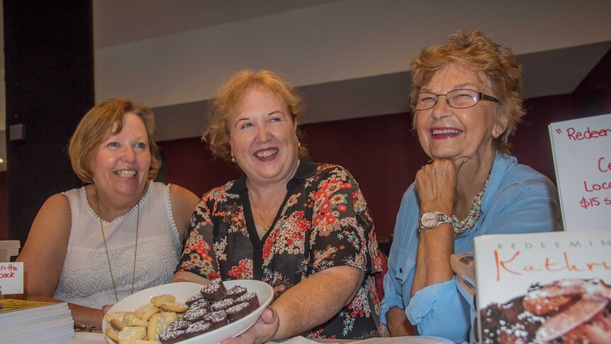 Three women laughing and smiling, one holding a heart shaped plate of biscuits sitting at a table