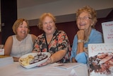 Three women laughing and smiling, one holding a heart shaped plate of biscuits sitting at a table