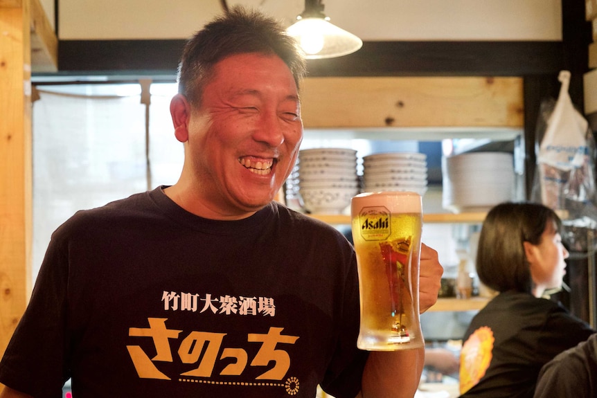 Masashi Sano smiles broadly while holding up a large glass of beer.