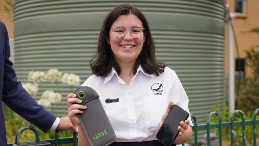 A girl wearing a white shirt and glasses smiles while holding a grey pouch in one hand and a phone in the other