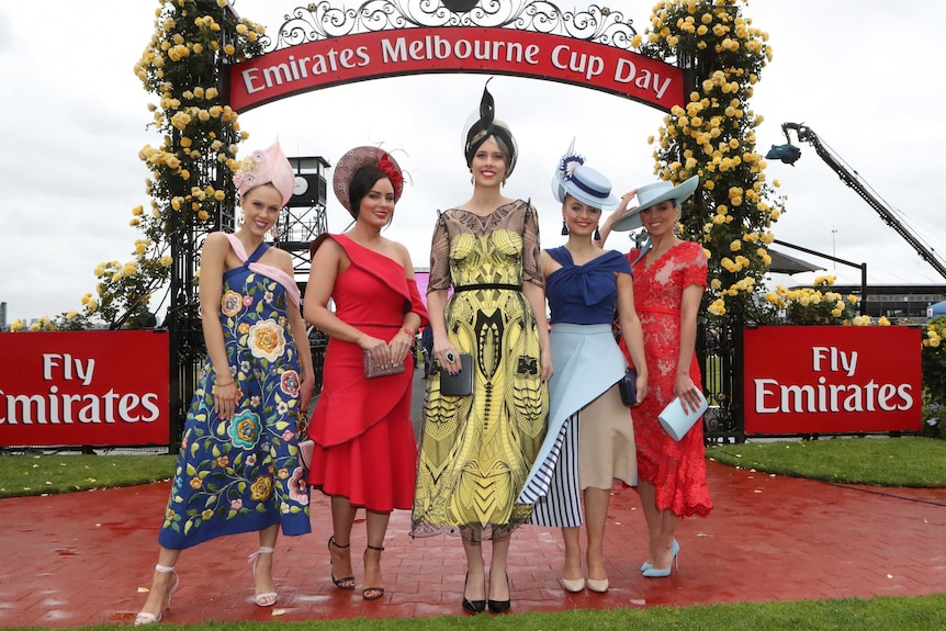 Four women wear colourful dresses in front of a Melbourne Cup day sign.
