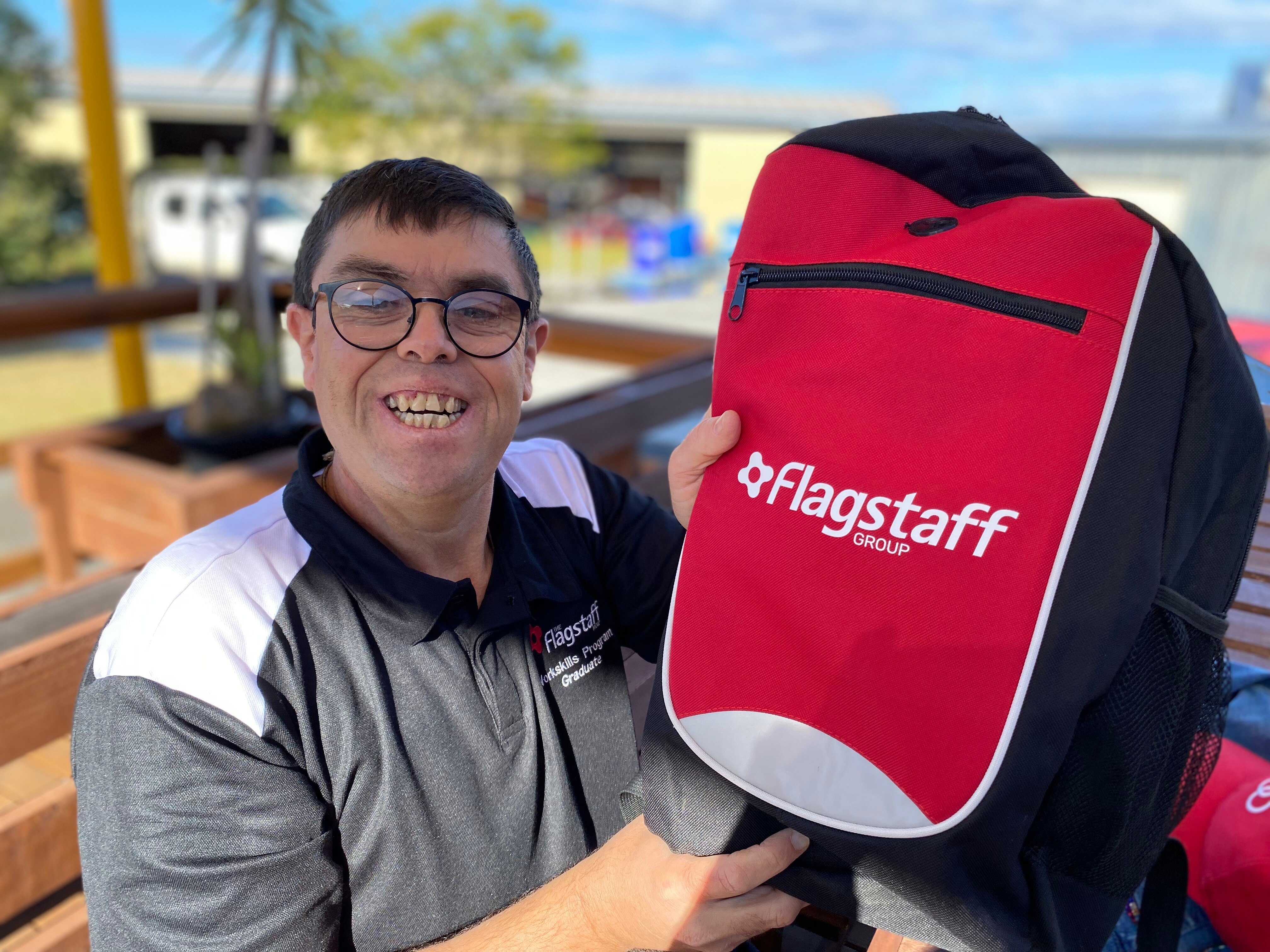 A widely smiling man with dark hair, glasses, wears white and grey t-shirt, Flagstaff logo, holds up a red backpack.