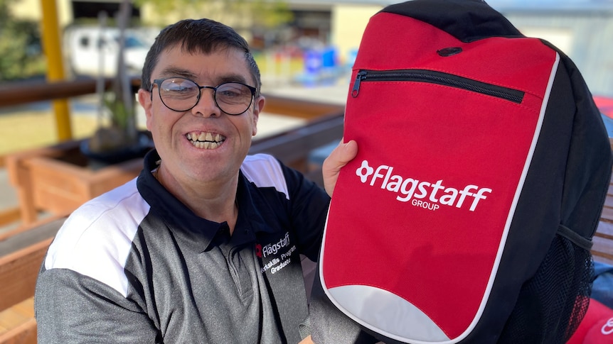 A widely smiling man with dark hair, glasses, wears white and grey t-shirt, Flagstaff logo, holds up a red backpack.