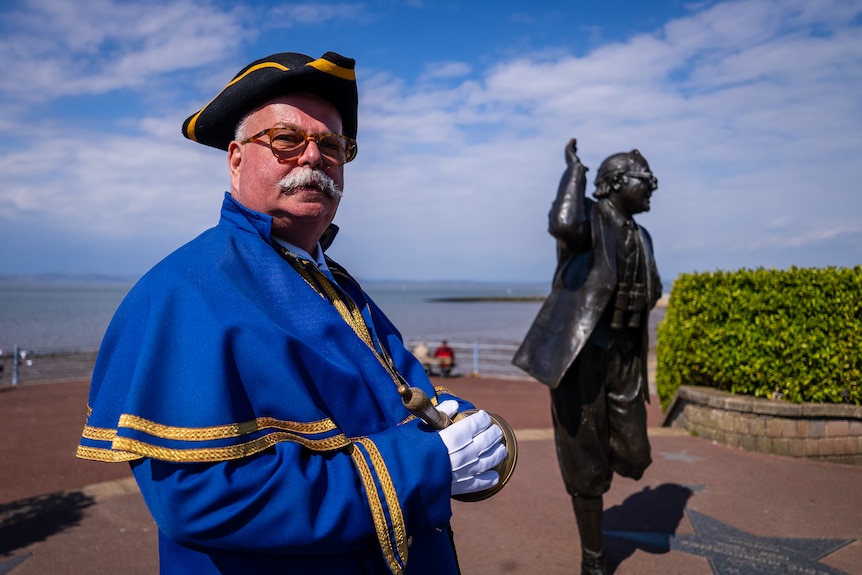 Town crier dressed in a blue robe with yellow details around the hem, standing next to a statue. 