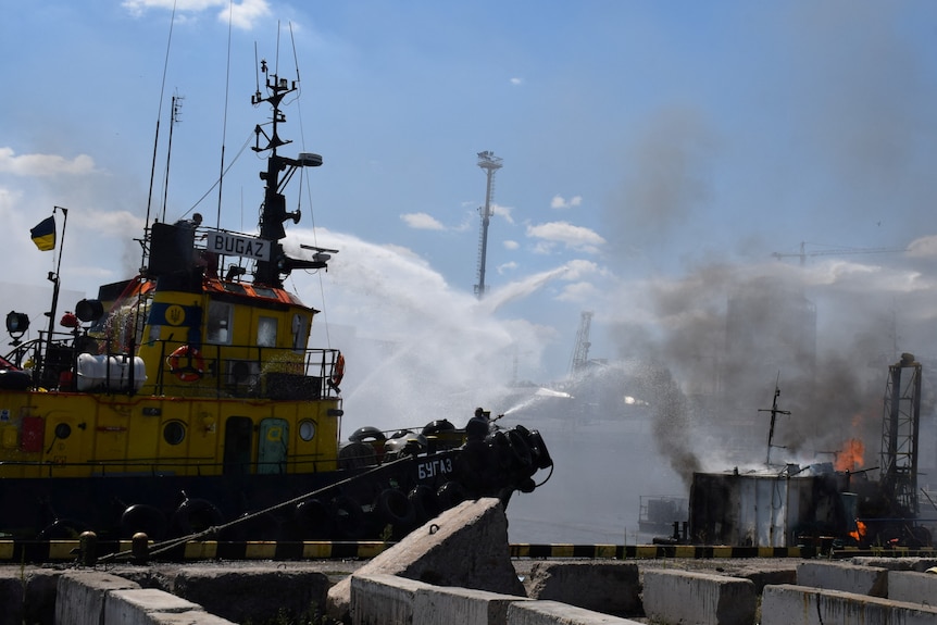 Firefighters work at site of fire at Odesa port, with ship seen in background as smoke rises nearby.