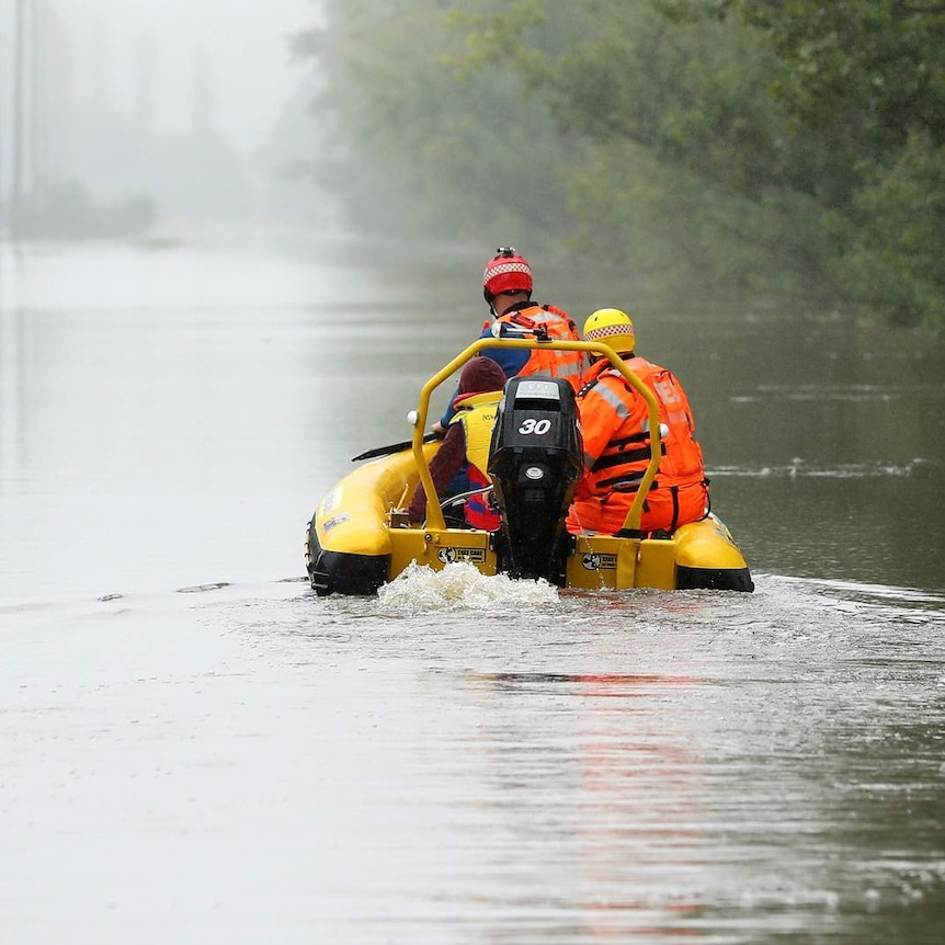 SES staff in orange and yellow gear sit on a water craft, churning through flooded waters. Water laps the bottom of a speed sign