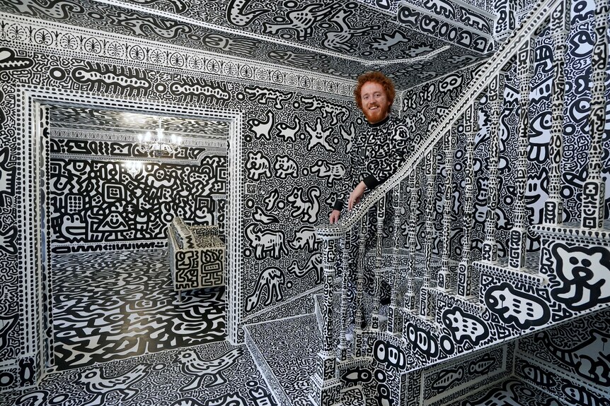 Sam Cox stands on the stairs of a room covered in black and white art. His clothes are also covered. 