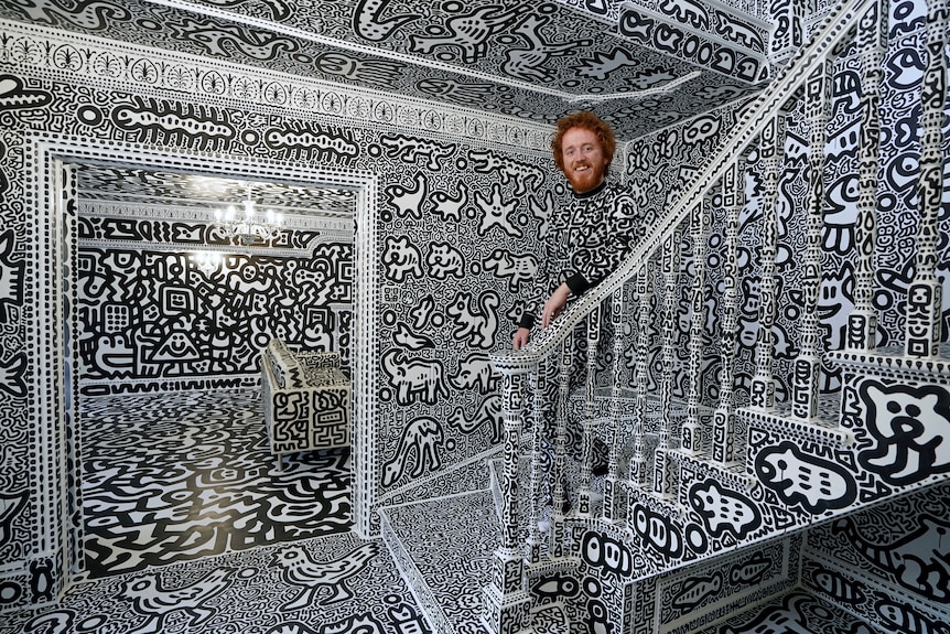 Sam Cox stands on the stairs of a room covered in black and white art. His clothes are also covered. 