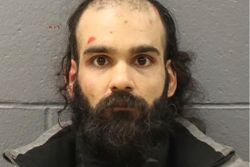 A mugshot taken against a brick wall shows a balding ban with a beard looking directly at the camera
