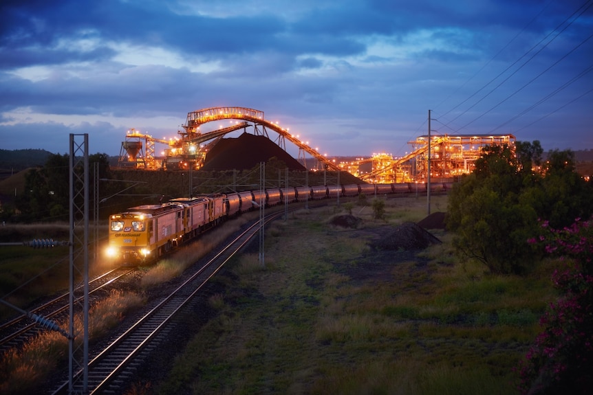 Long train with many carriages snakes its way along a track from a well-lit coal-loading facility at night