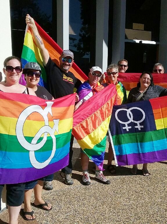 Eight people hold four rainbow flags outside SCRC council chamber at Nambour