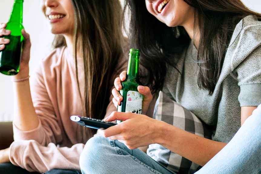 Close-up of two young women with brown hair laughing and drinking beer on the couch.